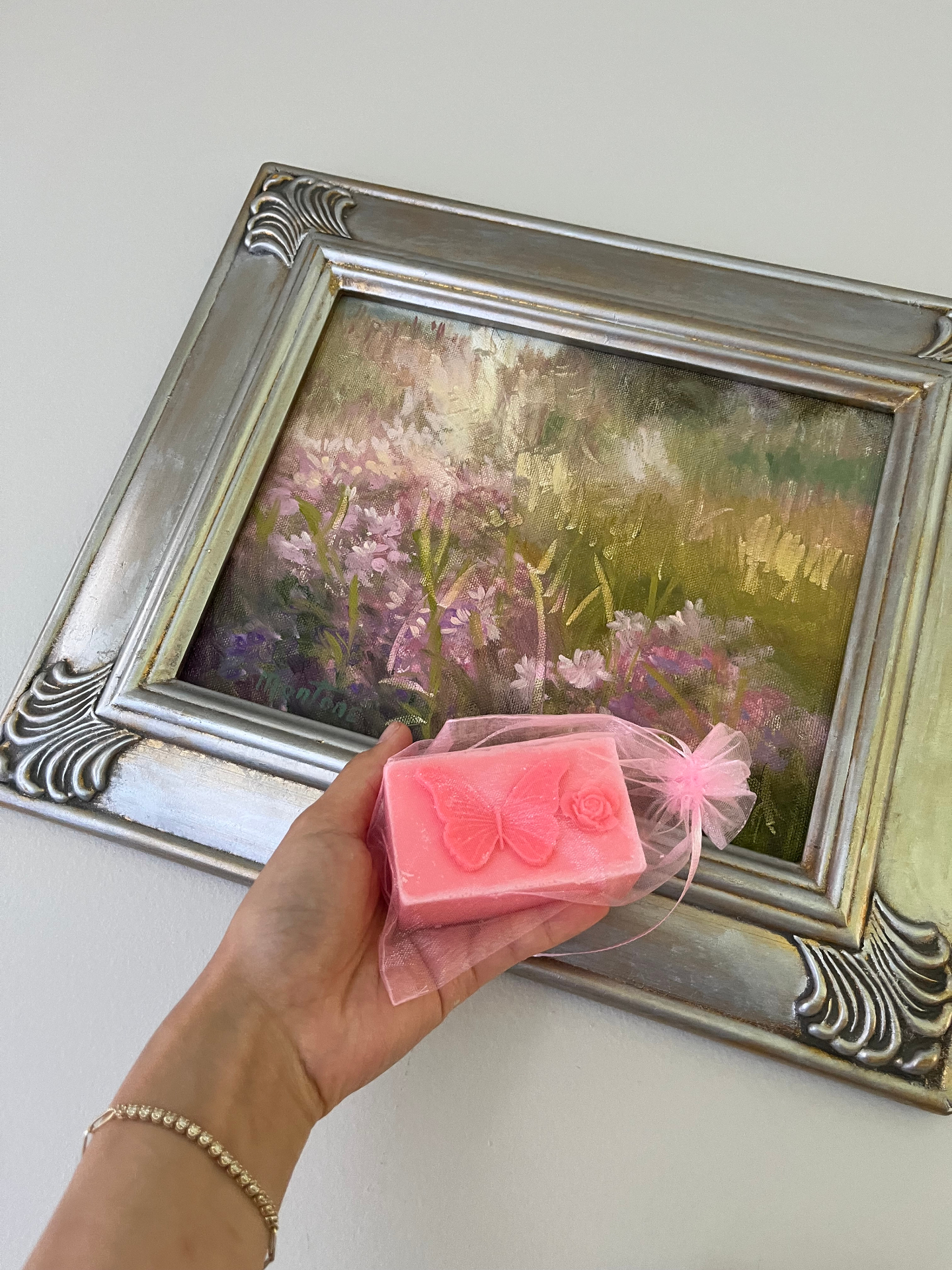 Pink butterfly soap in an organza pouch held against a silver-framed floral meadow painting