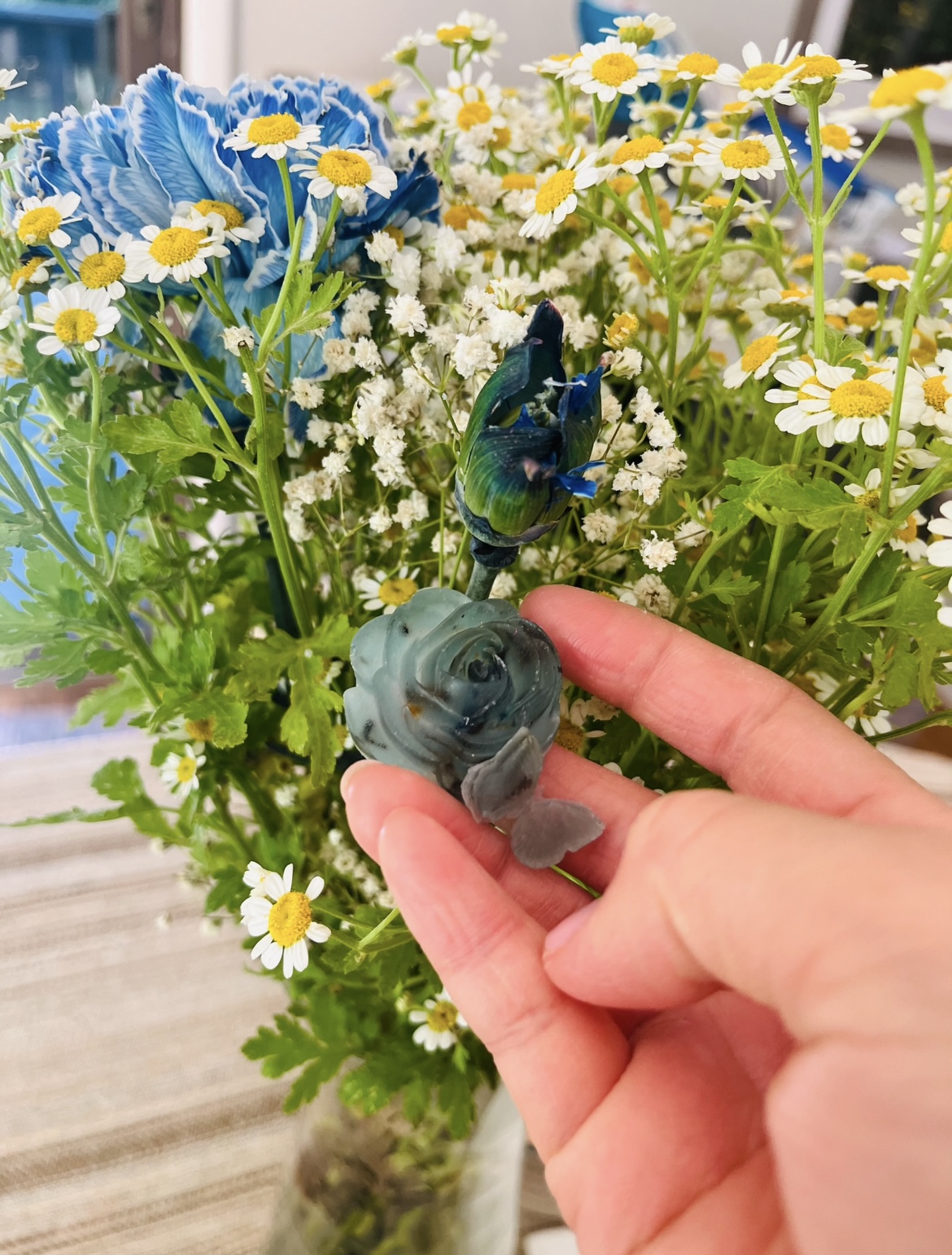 A hand holding a small blue rose soap among fresh daisies and blue blooms