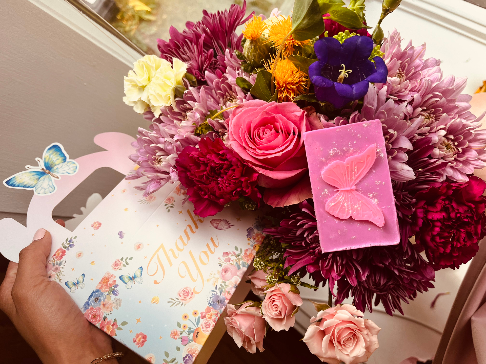 A pink butterfly soap inside a Thank You gift box, resting on a bouquet of pink, purple, and yellow flowers